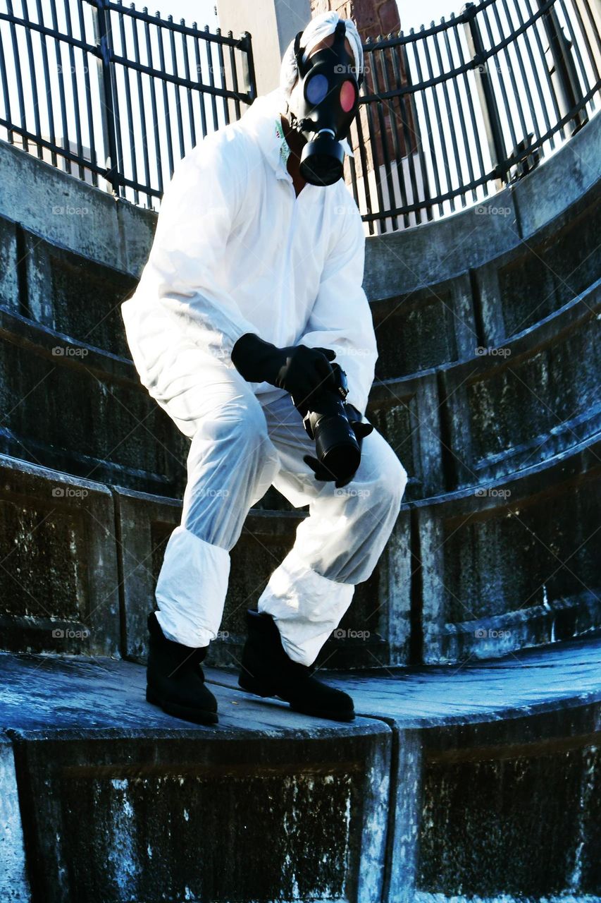 A man fully clothed from head to toe in a biohazard suit wearing a tactical gas mask brandishes a camera with a long lens.