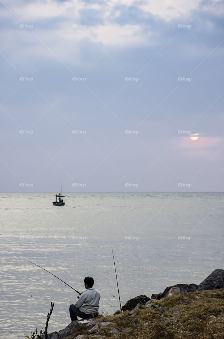 A man holding a fishing rod on the rock by the sea and a morning sunrise.