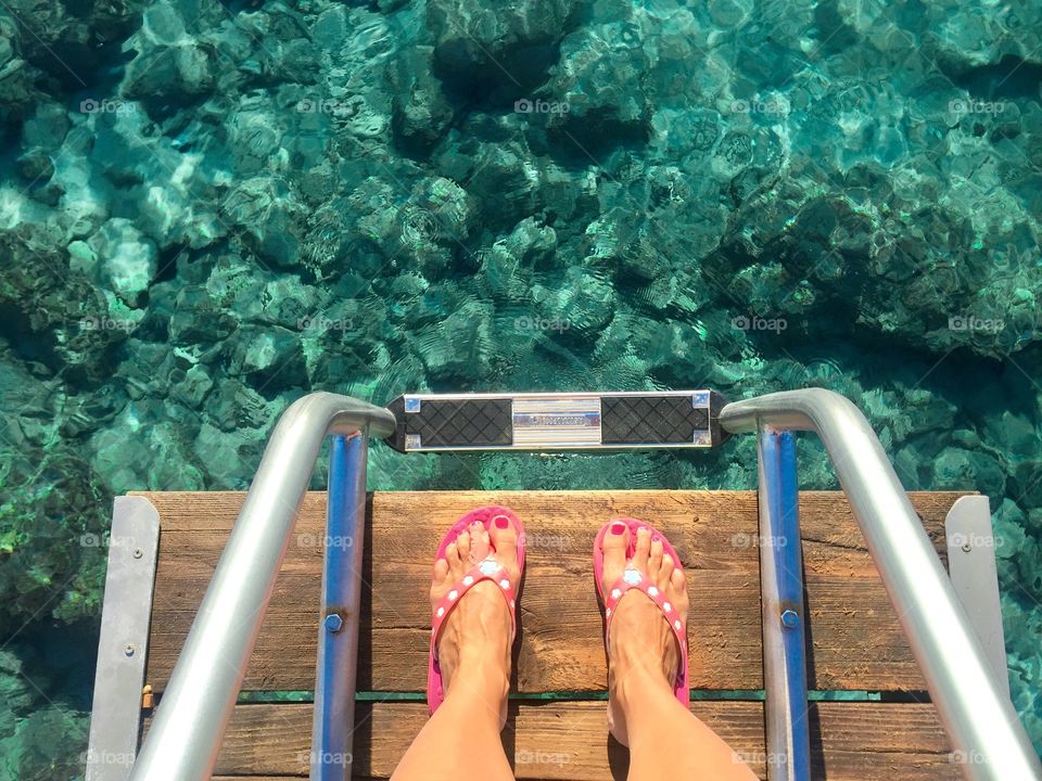 Woman s feet wearing pink flip flops standing near turquoise sea water in Greece