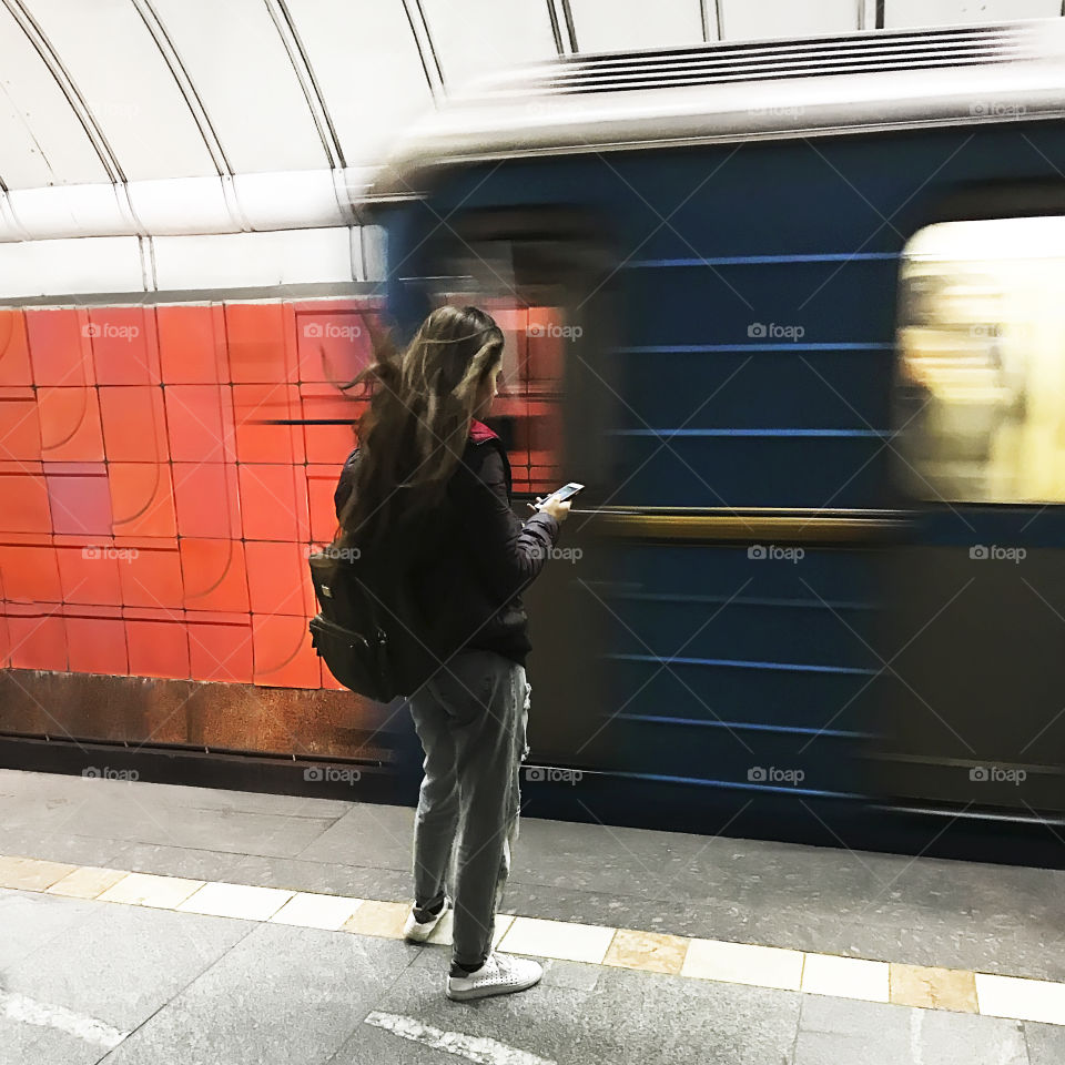 Young woman with a backpack using mobile phone while waiting for a train at city subway station 