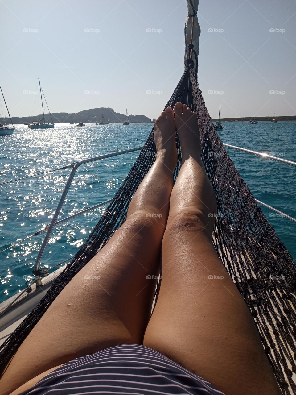 Female tanned Legs and Feet in a reclining position in a Black Net Hammock on the Bough of a Yacht looking out across the Ocean at La Pelosa Anchorage in Sassari Province, Sardinia, Italy. The Blue Sea is shimmering and glistening like Stars