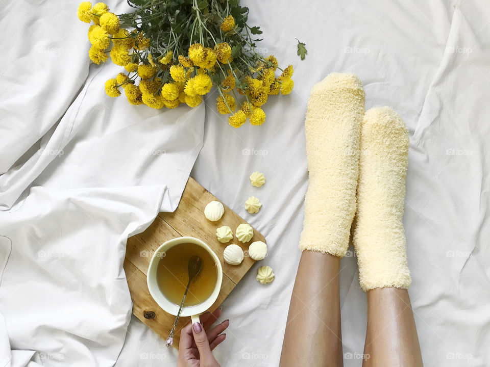 Young woman wearing cozy yellow socks and drinking tea with yellow candies in cozy bed with yellow flowers 
