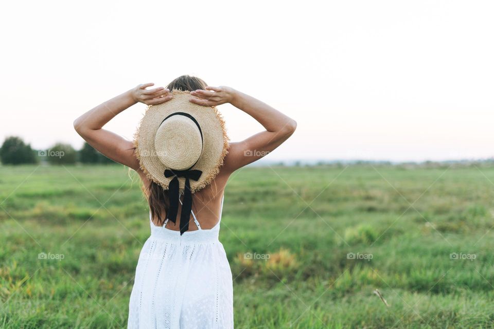 Young beautiful carefree long hair woman in white dress with straw hat in sunset field, view from back. Sensitivity to nature concept