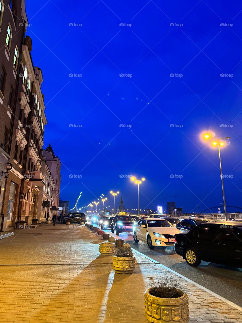 Ночная улица окутана мягким полумраком,освещенным фонарями,бросающими желтоватый свет. Машины медленно двигаются по дороге, их фонари бросают яркие лучи вперед, освещая асфальт и окружающие здания 