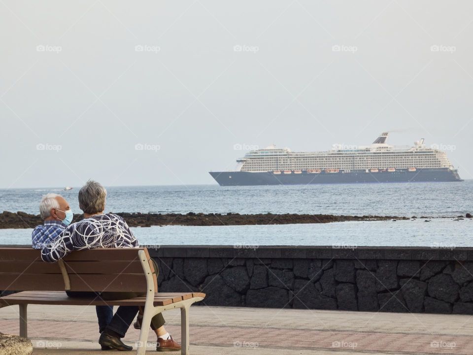 retired senior couple resting on a bench on a promenade overlooking the sea while a cruise ship passes by