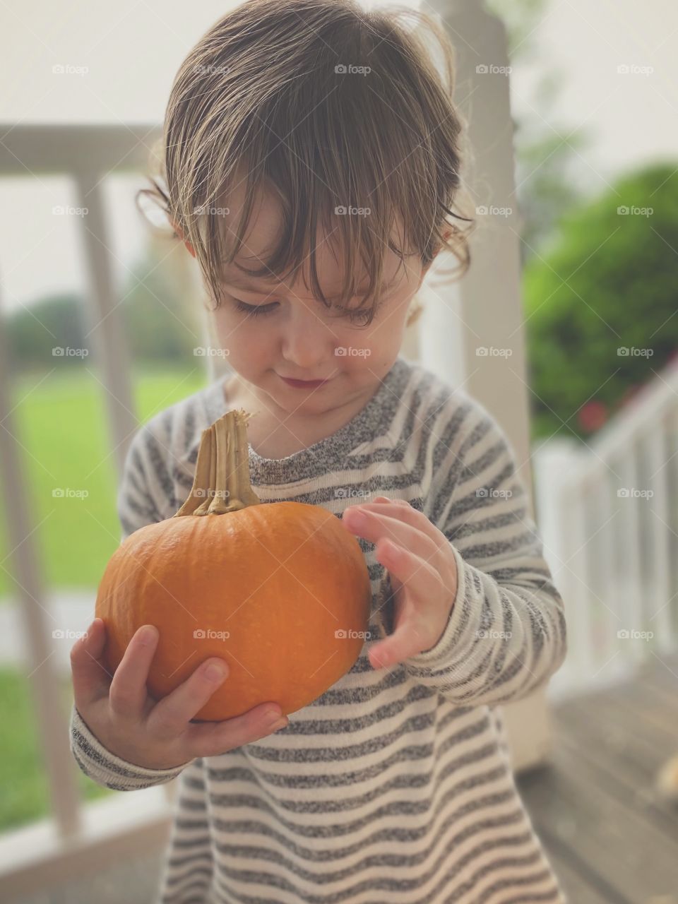 A curious young girl holds a small, bright orange pumpkin outside as she inspects it’s texture