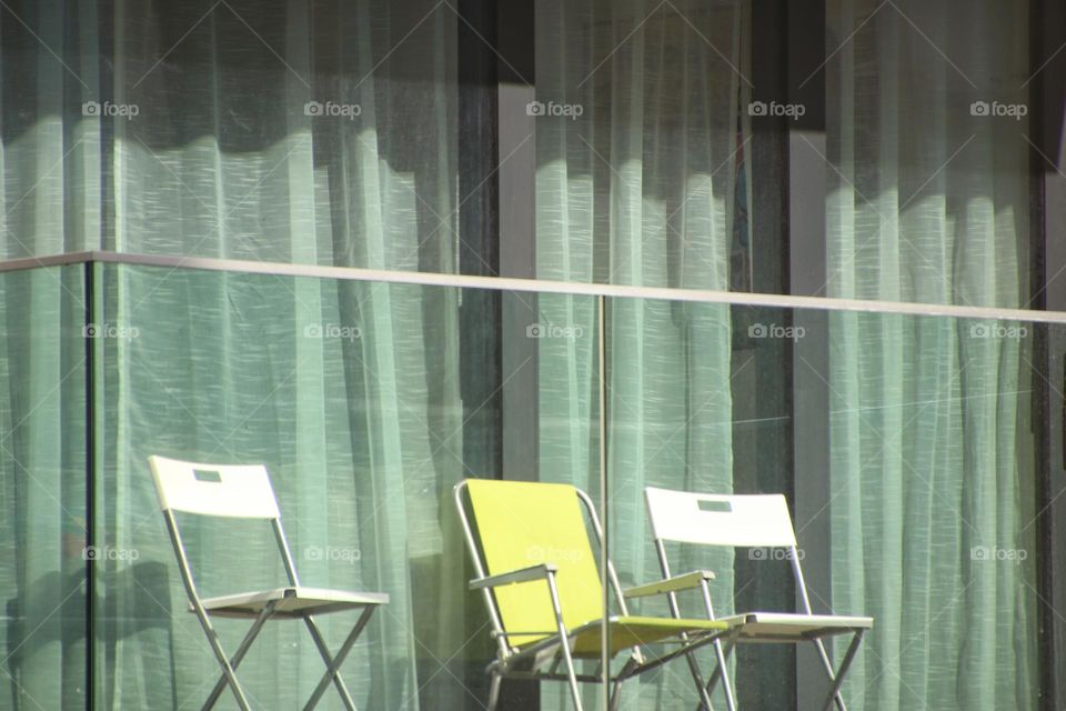 Three empty chairs are on a balcony in front of a curtained window