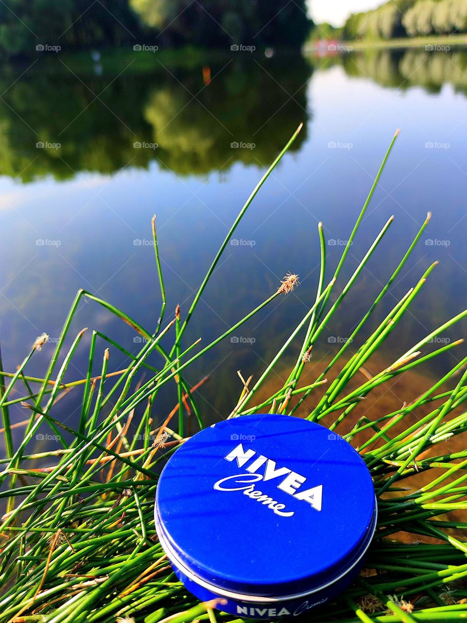 On the green grass of the river bank lies a blue box with the inscription: "Nivea cream". In the background, trees and a blue sky with white clouds are reflected in the water.