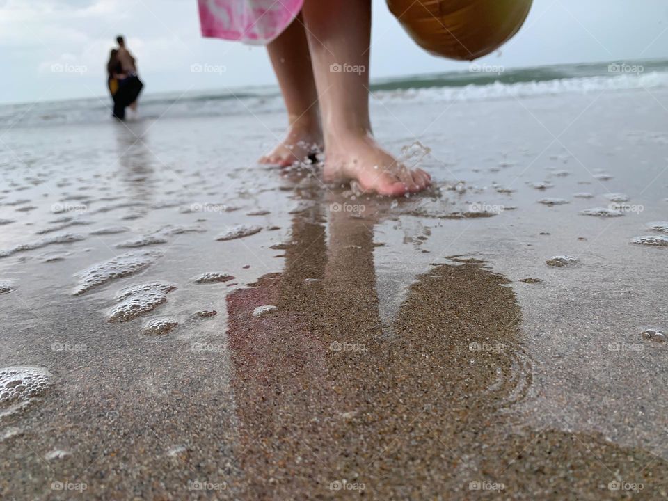 At The Ocean Seashore Beach With Girl Child Standing On The Wet Sand From Left Over Waves With Reflection And Another Person Further Walking In The Waves.