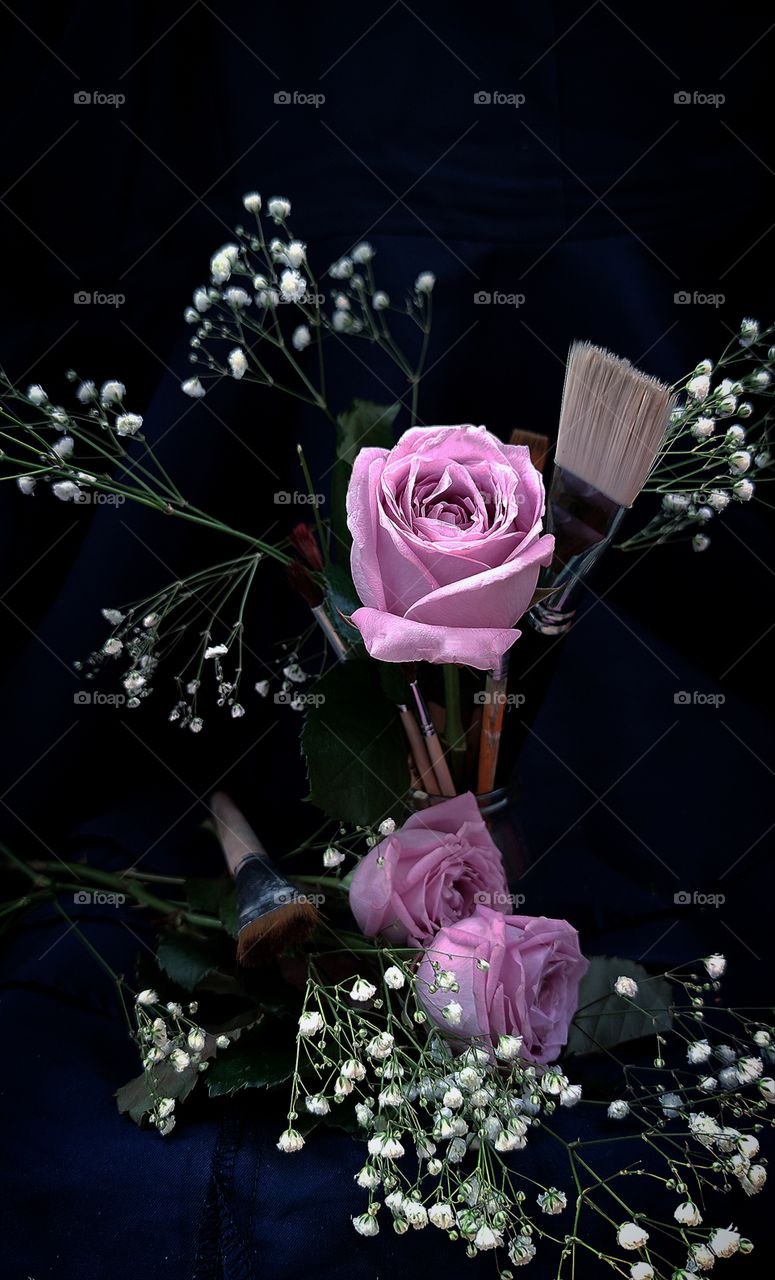 A purple rose stands with brushes for paints and white small flowers.  Two purple roses lie next to a paint brush and with white small flowers.  Background dark blue