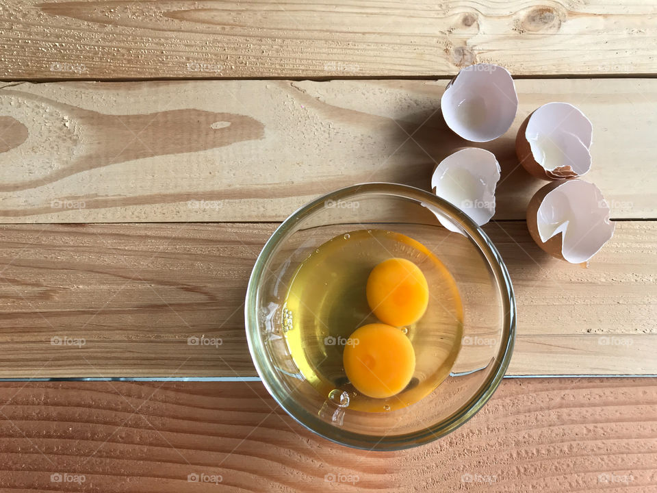 two fresh yolk and egg white in glass cup and eggshells beside it on natural rubber wood board with copy space on left side of frame