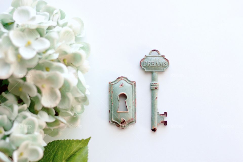 Overhead view of light teal, vintage, metal key with embossed word, “Dreams,” and keyhole with light teal hydrangeas on white background - landscape