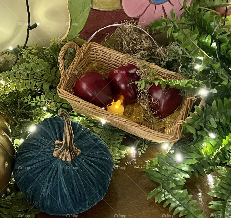 A small basket filled with apples resting on greenery with soft white lights next to a green pumpkin