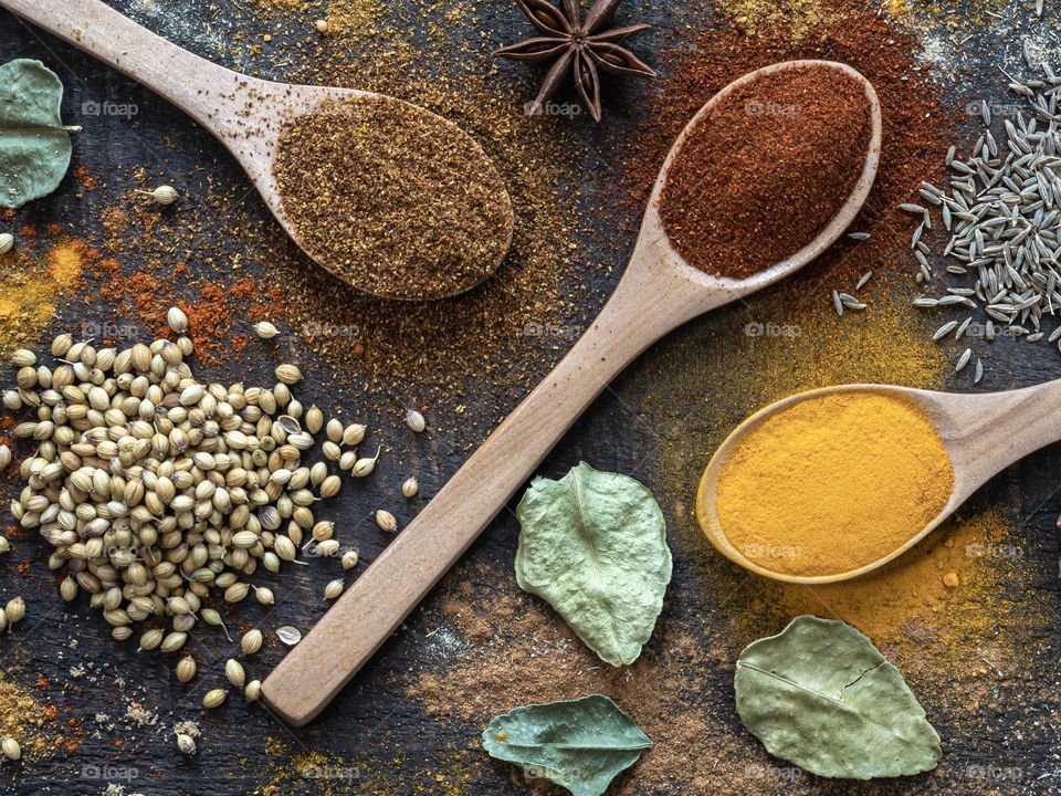 A dark wood background with spices in spoons, seeds & leaves 