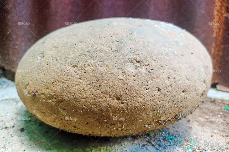 Close-up of a stone that is oval in shape and has a rough texture, placed near a brown wall. The stone looks brownish with a little dust or dirt stuck to its surface