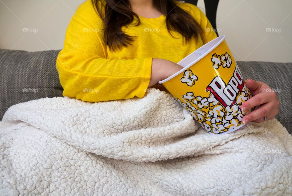 A young girl in a yellow T-shirt on a sofa with a bucket of popcorn covers in a soft blanket.