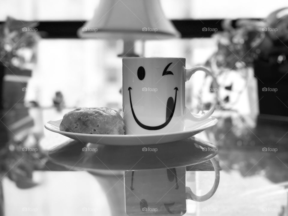 Reflection of a white cup with smiley design. Reflection of plate, leaves and lamp in a glass table. black and white.