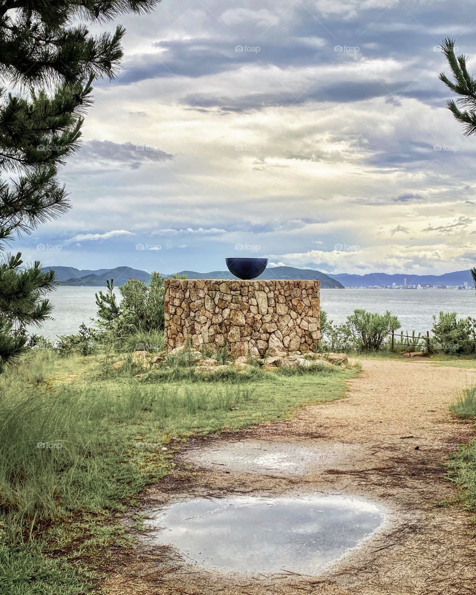 Puddles, clouds and outdoor art installation on Naoshima Island on a rainy day, with a seascape backdrop and muted tones.