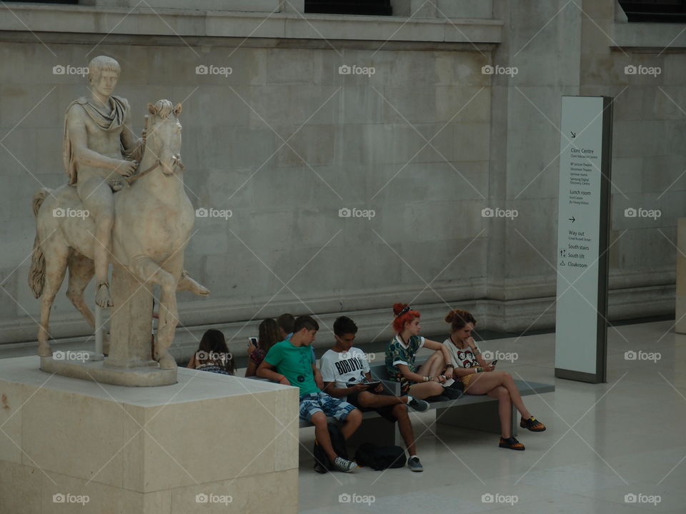 A group of young people sitting against a stone wall next to a statue in a lobby. 