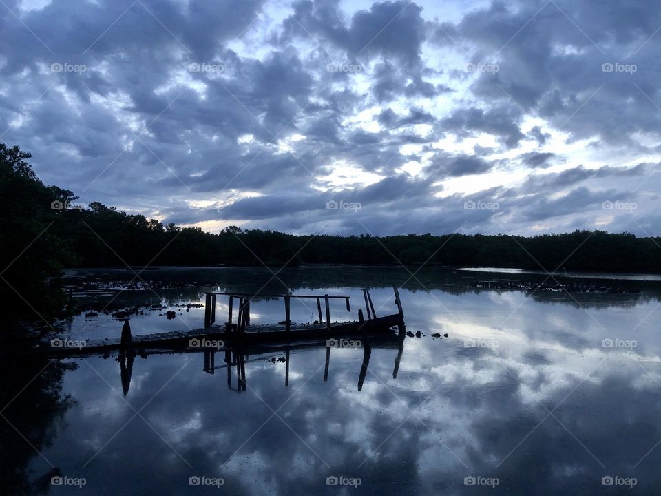 Dawn skies streaked with clouds reflect over the calm Bayou. Silhouettes of an old pier and shoreline are the only things breaking up the scene.