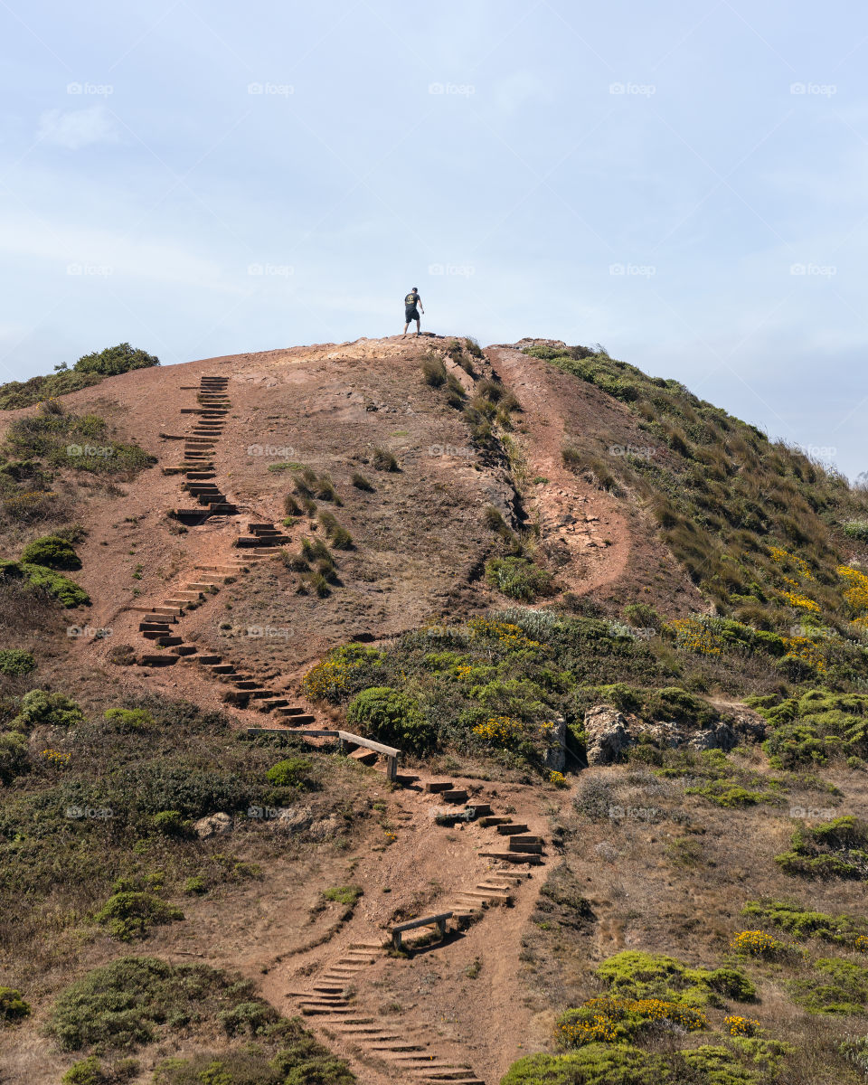 Lone person walking up a trail with long stone staircase switchbacks to the summit of a tall hill