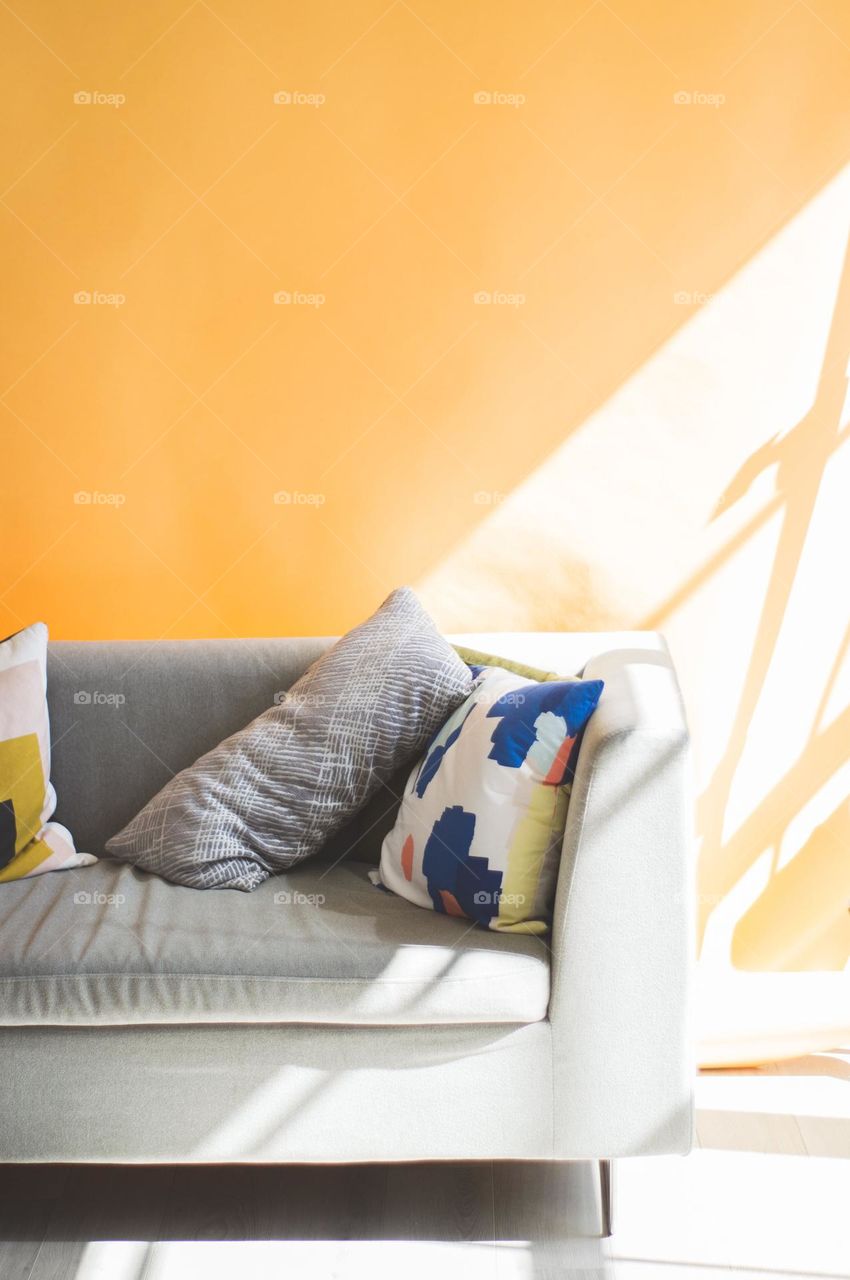 "The sun shines into the room with yellow walls, making it even brighter.
Light falls on a gray modern style sofa with some colorful cushions."