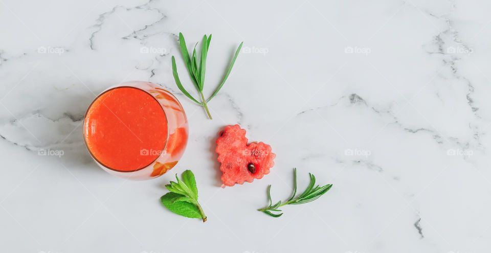 Natural watermelon drink in a glass with a heart-shaped watermelon slice and mint leaves with rosemary sprigs lie on the left on a light concrete background with copy space on the right, close-up flat lay. Summer drinks concept.