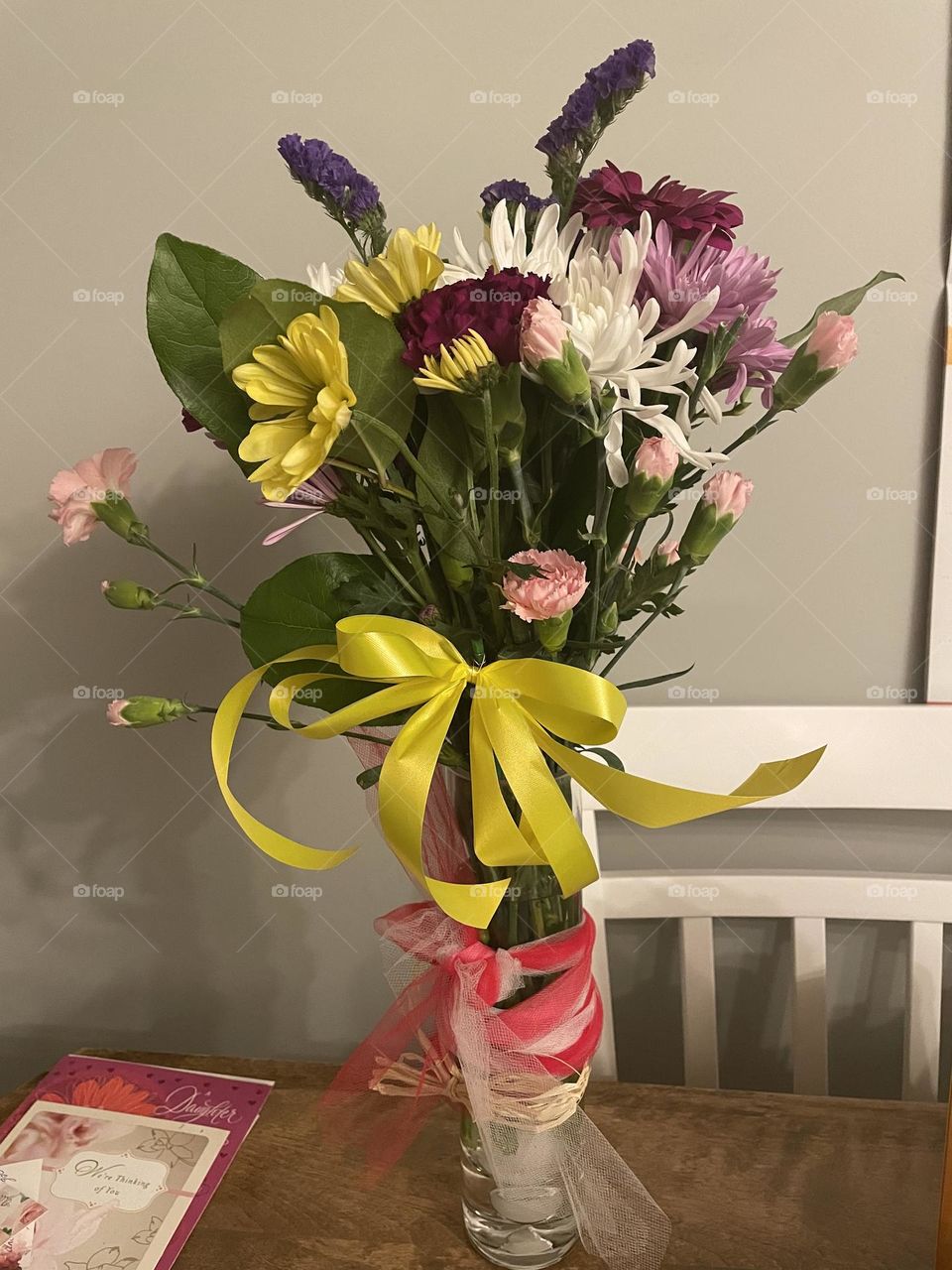 Flowers in a glass vase, with yellow ribbon and a red and white ribbon, that I also got when my youngest son passed away, 2/22/22, he was only 34, from an OD, from my mom and dad, card also laying on table.