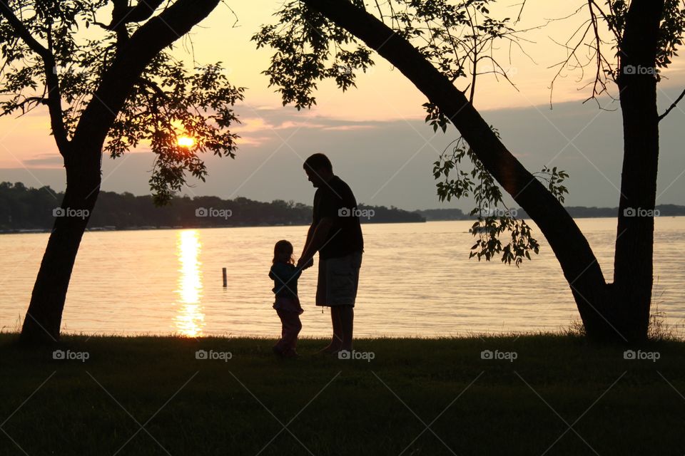 Gorgeous golden bright yellow sun is setting off in the distant horizon with beautiful silhouette of father and daughter holding hands under trees! 