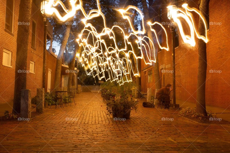 Exposed brick road at nighttime with trees lit up with bright lights. Brick buildings surrounding exposed brick road. Old town  in New England 