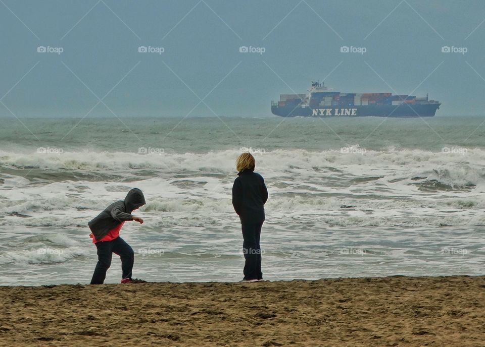 Boys Playing On A Beach With Cargo Ship In Background
