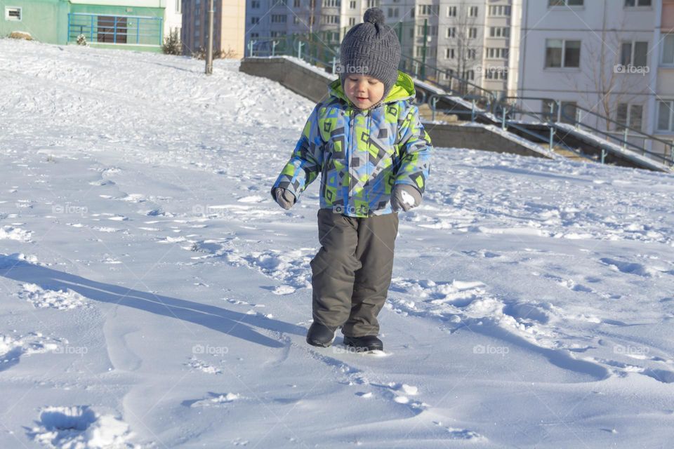 A child in winter in winter jackets, pants, a hat and boots on white snow on the street and in the park in nature plays winter fun and sleds.