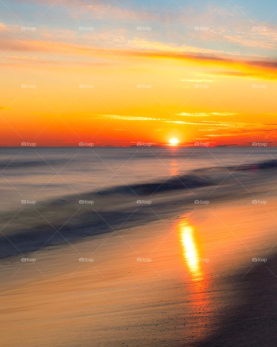 Sun setting on the horizon over the ocean with soft dreamy waves rolling in - reflection on sand