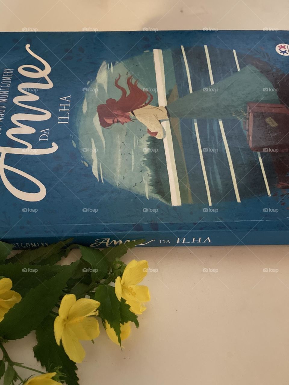Mesa branca com um livro de leitura e ao lado buquê de flores amarelas enfeitando a cena.