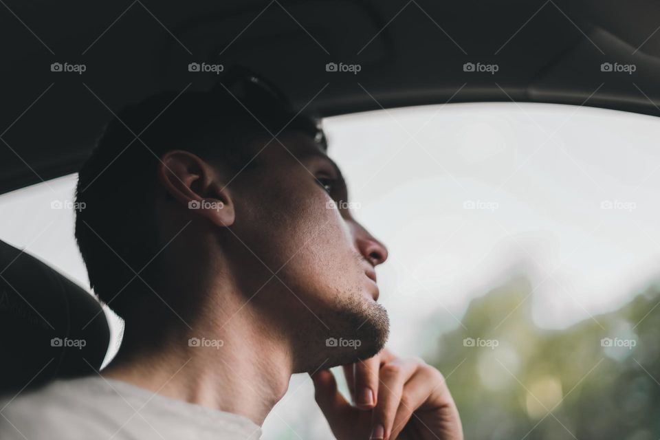 Portrait of a young handsome Caucasian guy sitting in his car and looking at the road with a fence, close-up view from the side. Road Trip concept.