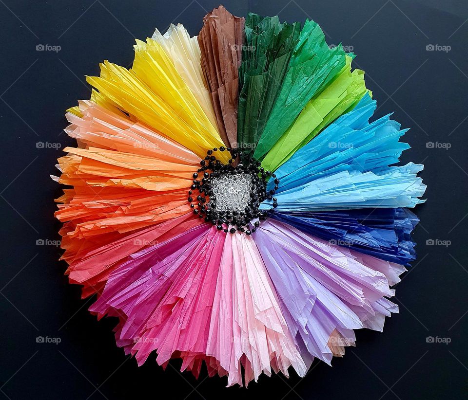 Flatlay composition of a multi-coloured flower made of silk papers, black and clear beads placed on black paper, creating a colour explosion