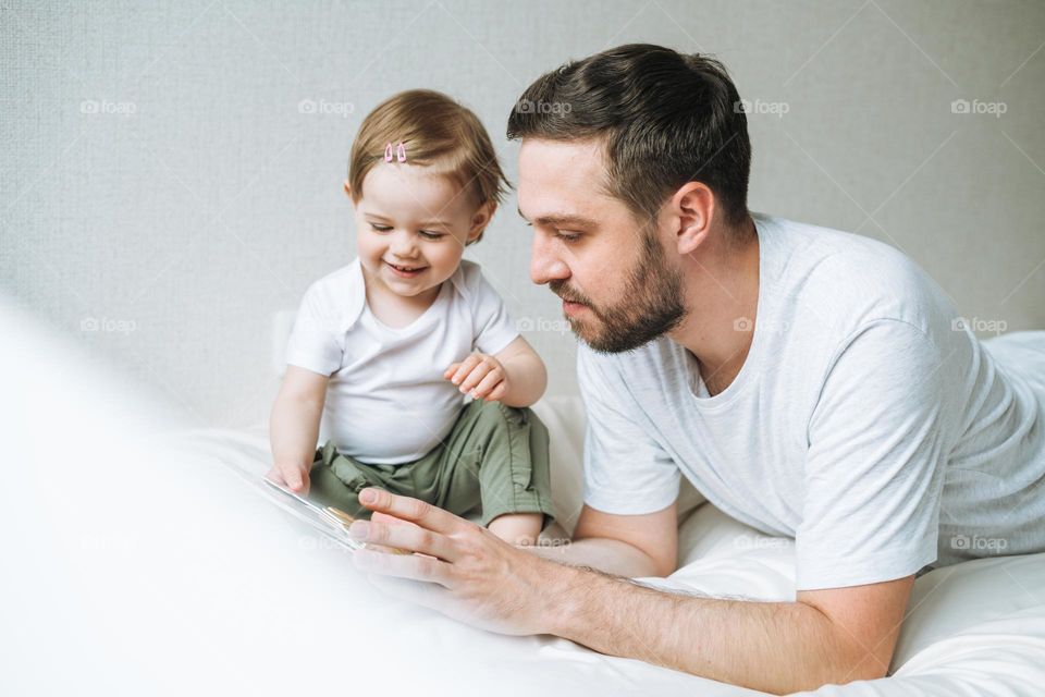 Happy father young man and baby girl little daughter having fun reading a book in children room at home