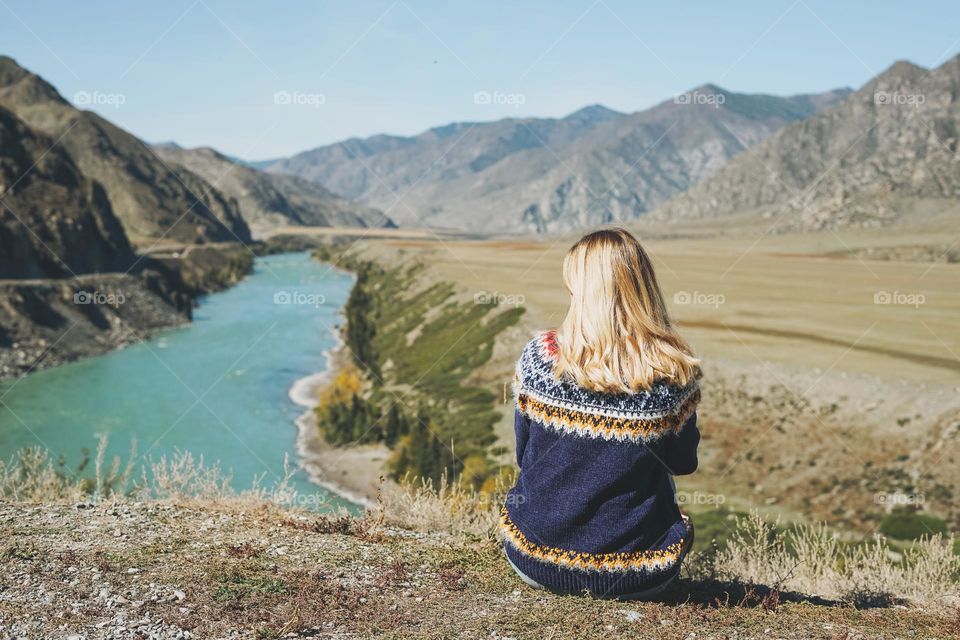 Young blonde woman in nordic sweater sitting on background of turquoise Katun river, Altai mountains