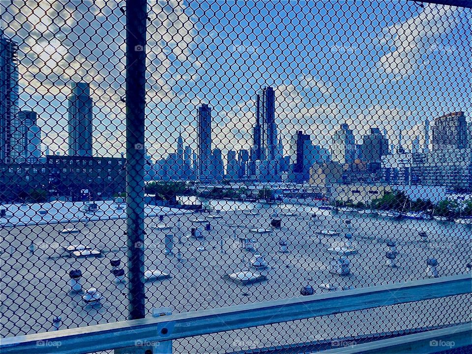 Looking thru a mesh fence on the “Greenpoint, Bklyn” side of the “Pulaski Bridge” at “Newtown Creek” at the “Manhattan” skyline, partly also silhouettes of “LIC, Queens” buildings behind the flat rooftop across the way. 2022. Hypnotic Productions