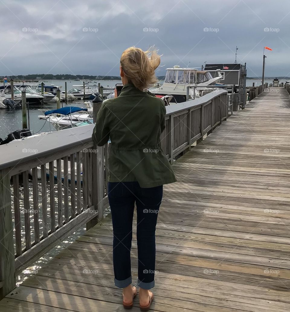 A woman is taking photos of bosts in a harbor with a cell phone while standing on a wooden pier to the ocean. 