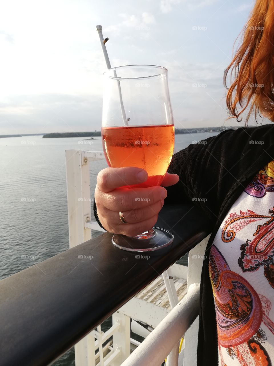 A red haired person is standing on the deck of a passenger ship and leaning on the black railing. She has an orange drink in her hand, a golden ring in her finger, wearing a jersey and a multicoloured shirt.