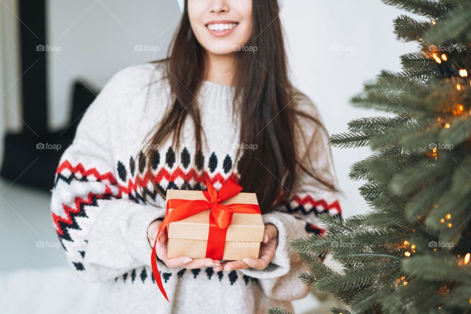 Young woman in cozy sweater with gift box with red ribbon in room with Christmas tree at home