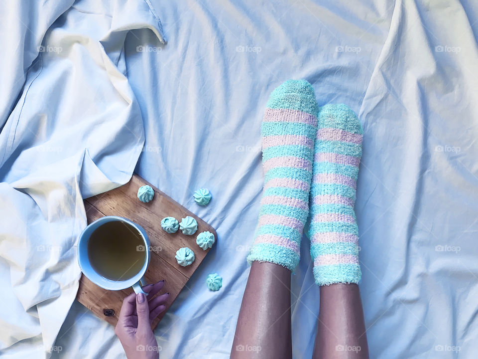 Young woman wearing cozy blue socks enjoying hot tea with candies in cozy bed 