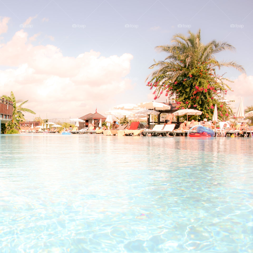 Großer Swimmingpool mit Sonnenschirmen und Palmen, blauer Himmel mit großen Wolken, Personen sind nicht zu erkennen aber vorhanden