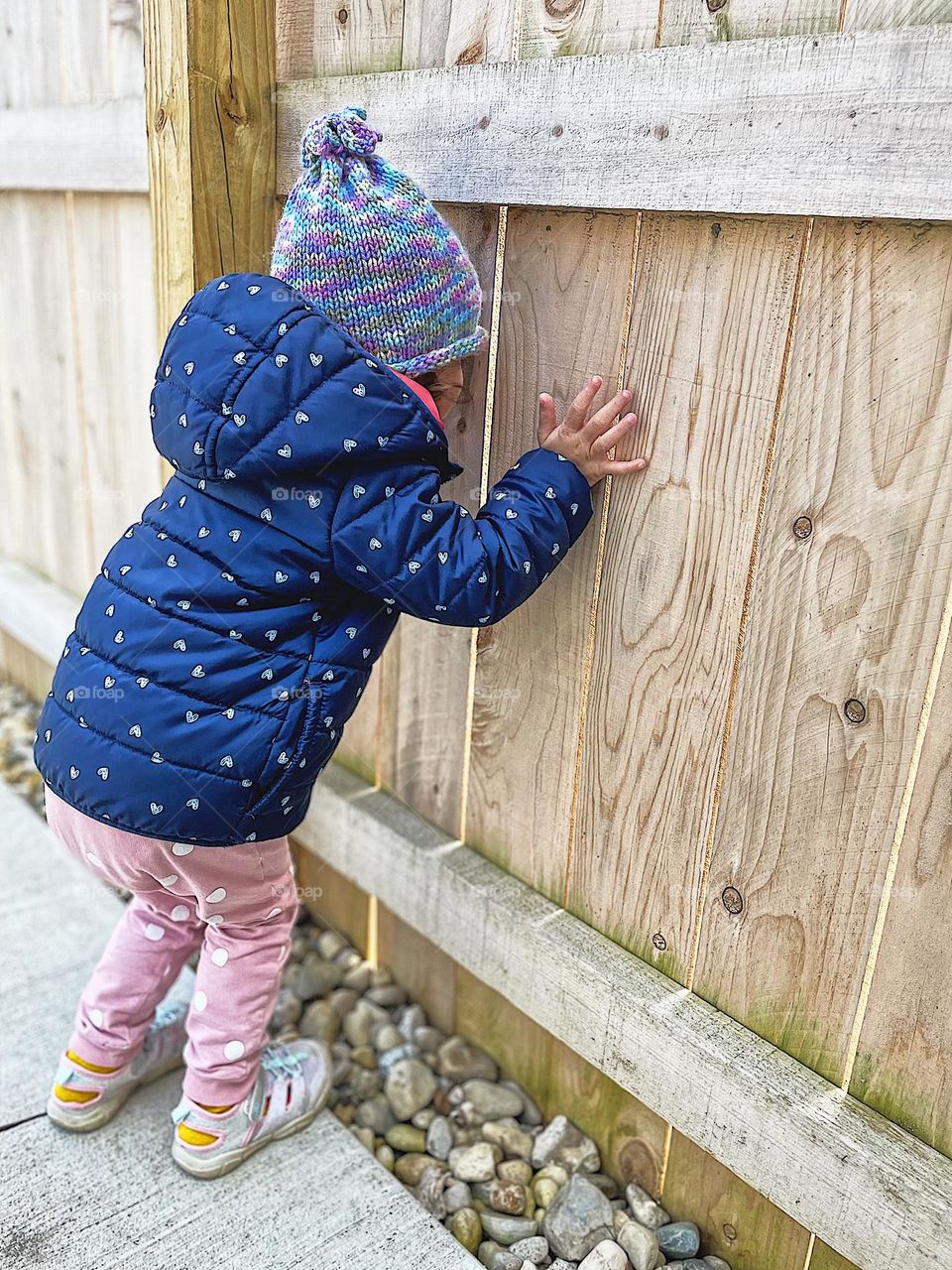 Child looks through crack in fence, toddler is fearful of what’s beyond the fence, fear of the unknown, curious toddler looks through crack in fence, looks to the other side, fear as an emotion in children, fearful child looks through crack in fence 