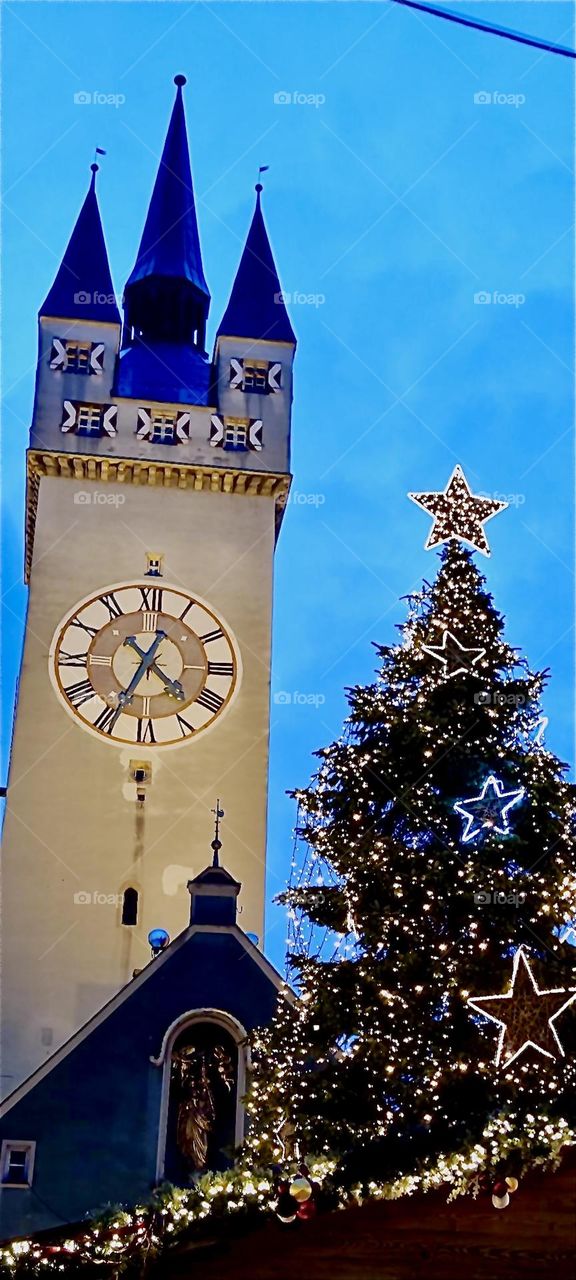 This is the medieval clock tower in the beautiful town of “Straubing”, “Bavaria”, Germany next to a very large illuminated Christmas tree to usher in Christmas and the holiday season. 2023. Hypnotic Productions