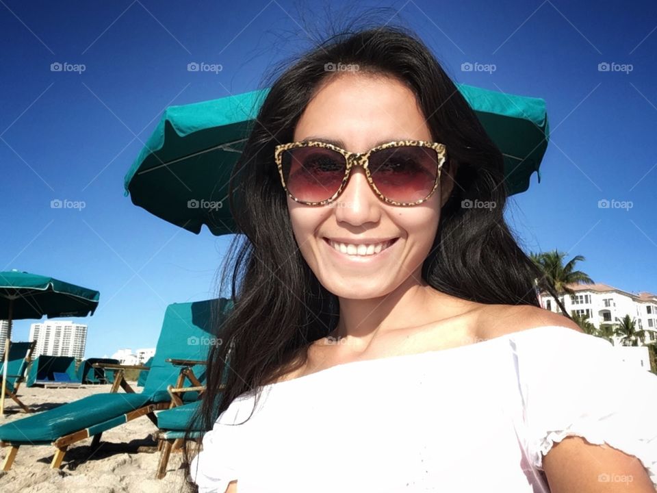 A woman smiling wearing leopard sunglasses with white dress in a blue sky background. In the beach with green beach chairs.