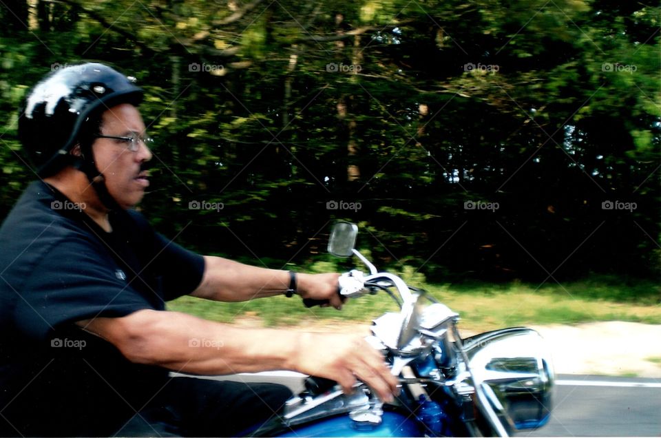 Man driving motorcycle on road wearing helmet, green background as a pic is taken as we bikers are riding as a group in the summer. Wearing eyewear, short sleeve tee shirt, black helmet. He's a biker.