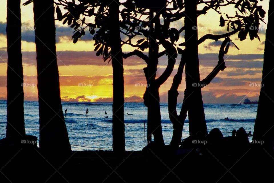 Sunset on Oahu’s Waikiki Beach. Unidentifiable surfers riding the waves. Clouds enhance the evening colors. 