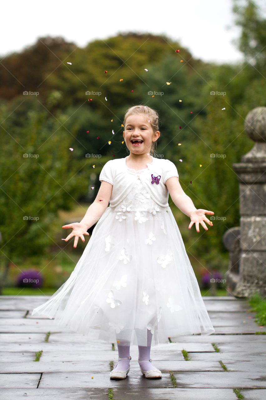 A young bridesmaid or flower girl in a white dress throwing confetti in the air at a wedding whilst smiling and laughing.